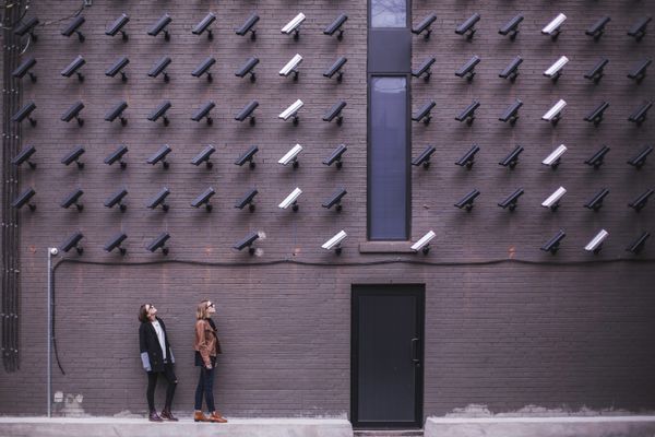 Dozens of surveillance cameras arrayed in a grid across a painted brick wall, two women on the pavement below looking up at them