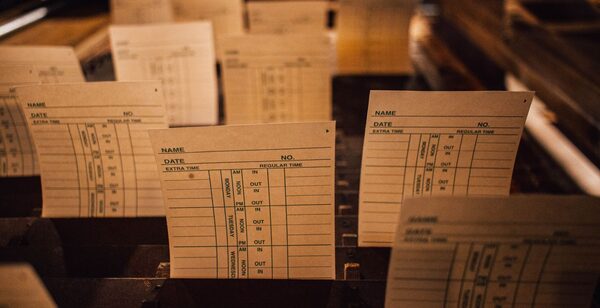 Blank paper time cards standing in a rack, each printed with NAME, DATE and columns headed IN and OUT