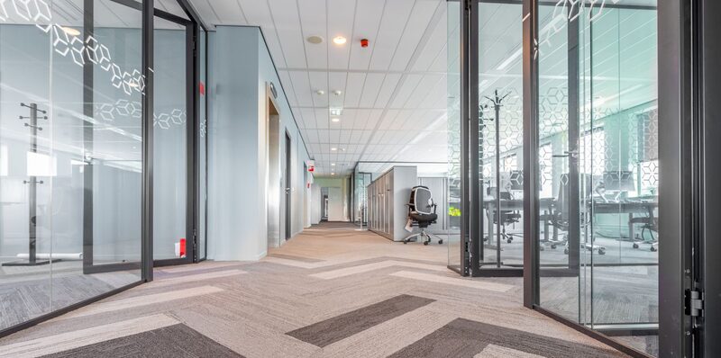 An office corridor lined with glass-partitioned meeting rooms, the partitions folded open onto empty desks and chairs